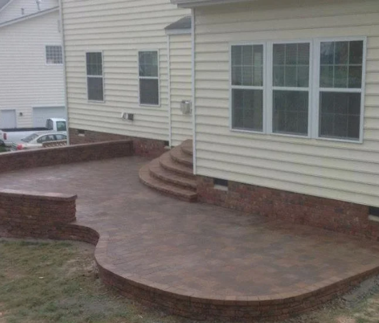 Curved brick patio beside a beige house with large windows and steps leading to a door.