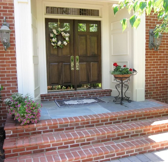 Brick front porch with wooden double doors, potted flowers, and three steps leading up to the entrance. 