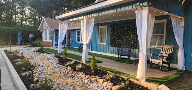 Blue cottage with white curtains, rocking chairs on porch, and a decorative garden in front.