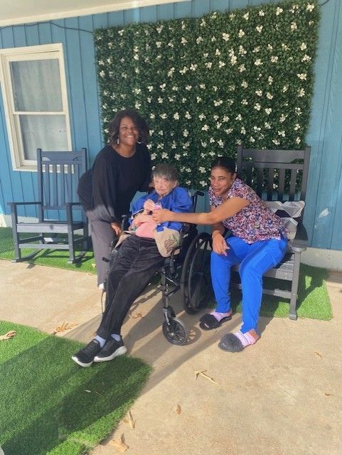 Three people pose outdoors: one in wheelchair, two others stand. Green floral wall in background.