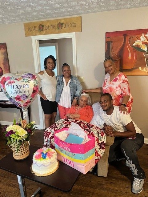 Five people gather for a birthday celebration; woman in a chair surrounded by gifts, cake, and balloons.