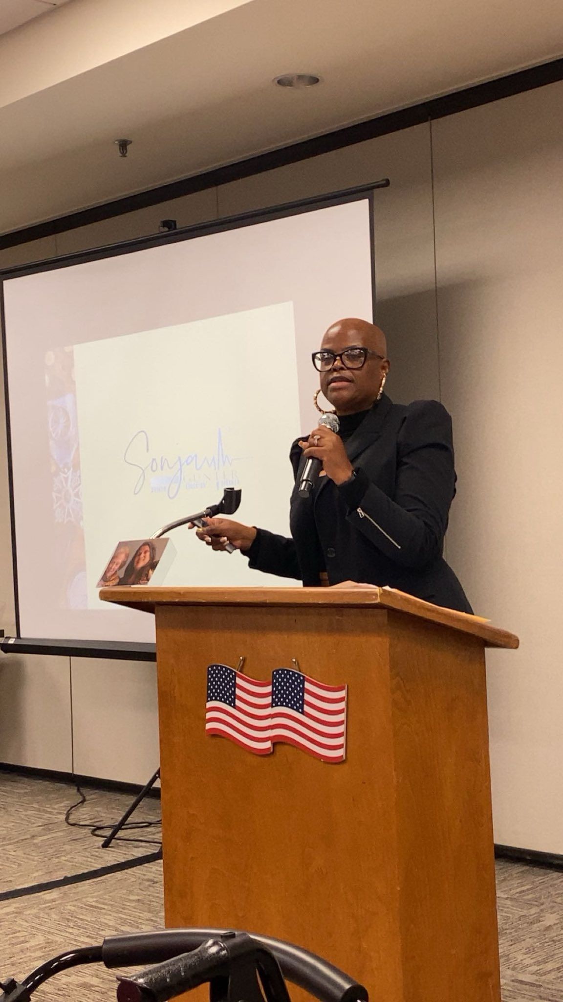 A person speaks at a podium with two American flag decals, addressing an audience in a room with a projection screen.