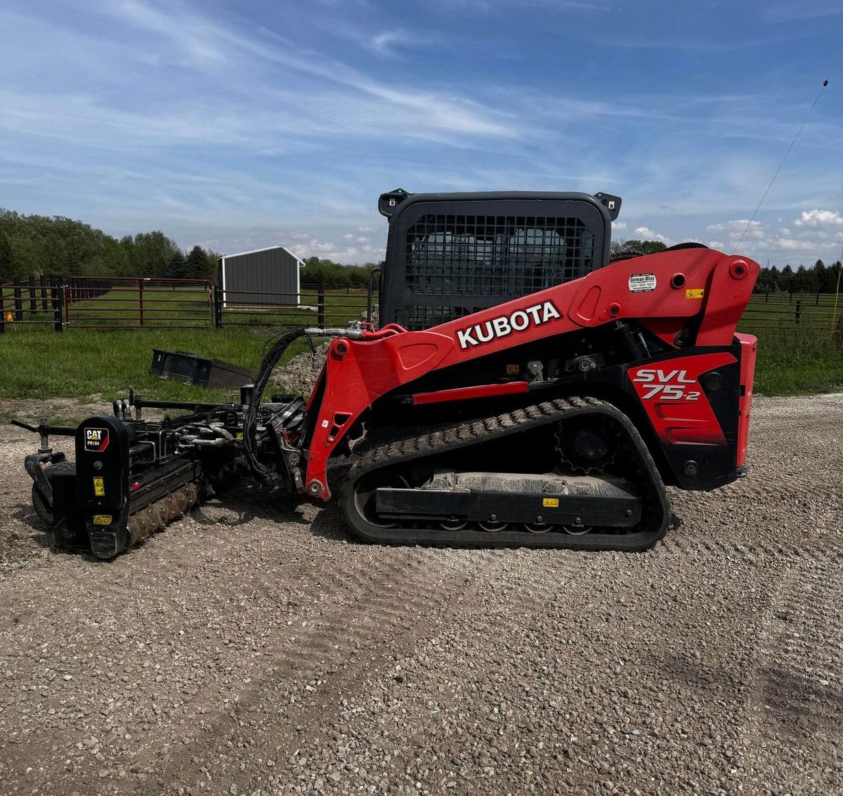 Red Kubota SVL 75 track skid steer with brush cutter attachment on gravel, field in background.