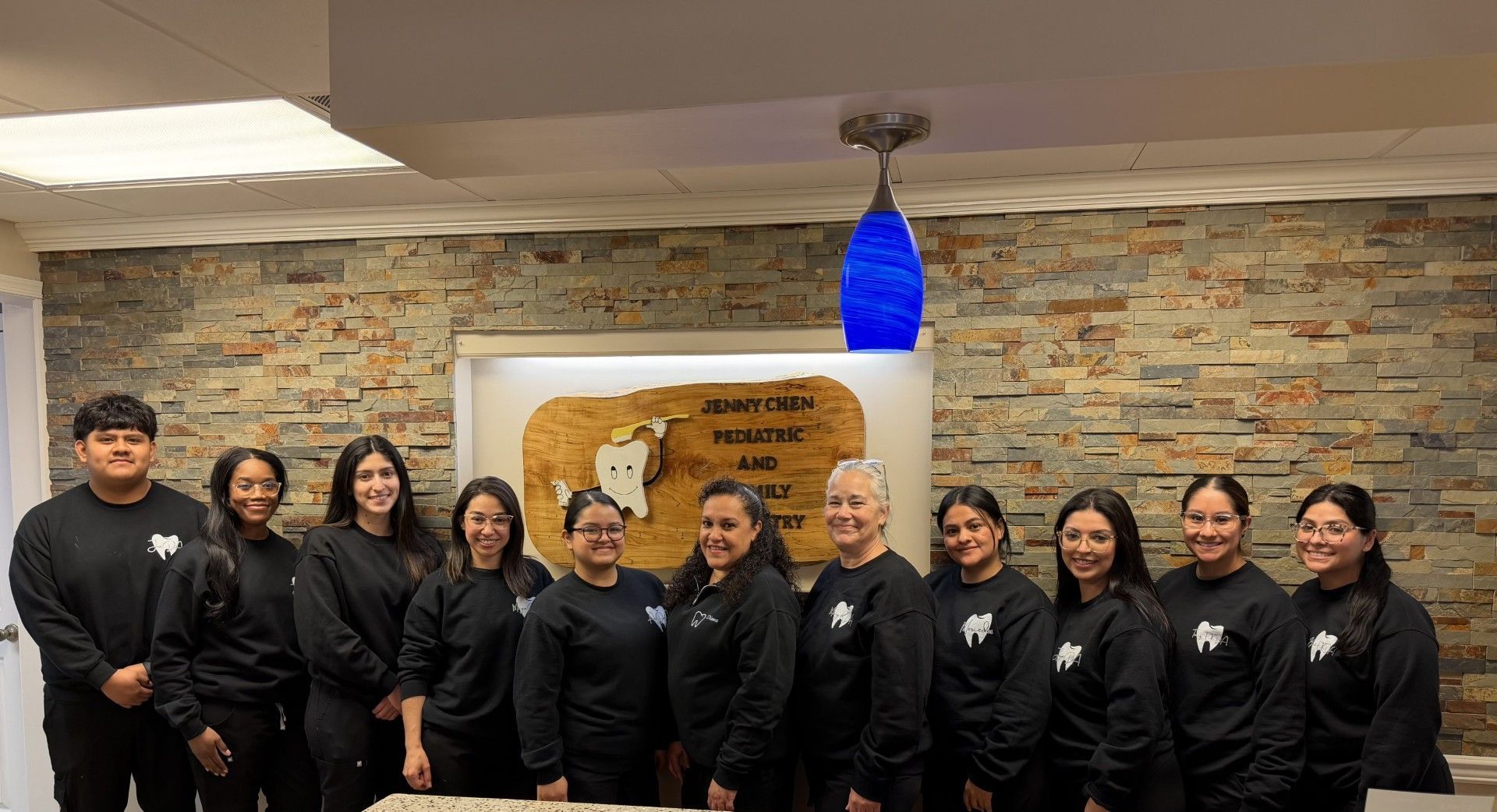 A group of eleven dental staff members wearing matching black sweatshirts stands in front of a stone wall in an office.