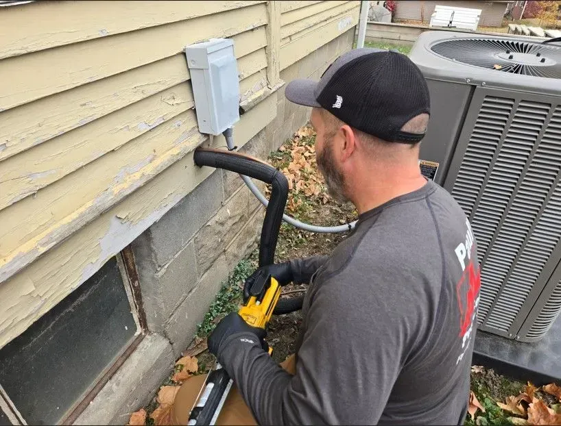 A person in a baseball cap works on HVAC tubing near a house with an air conditioning unit.