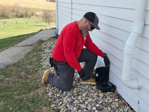Man kneeling beside a house, working on an outdoor HVAC unit near a downspout.