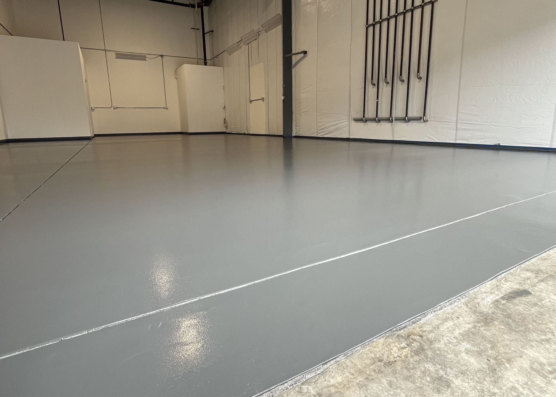 Empty gray-painted industrial space. Concrete floor is newly coated. White walls and overhead pipes are visible.