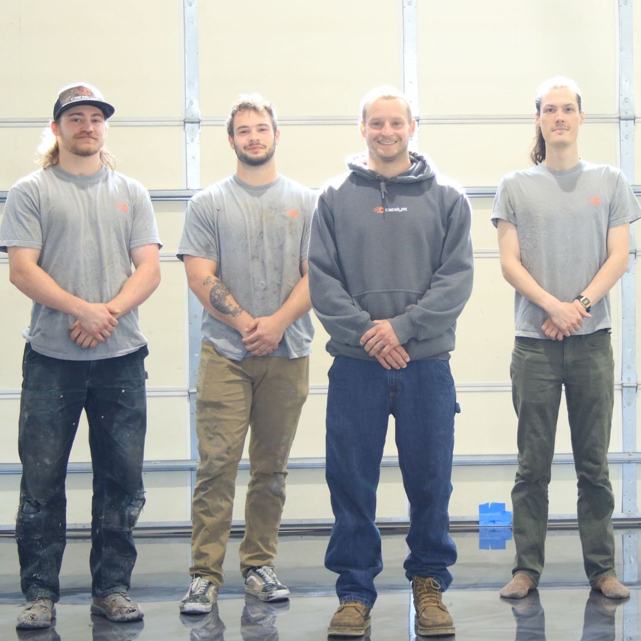 Four men stand in a garage-like setting, facing the camera. They wear work-related clothing; one has a hat, another has arm tattoos.
