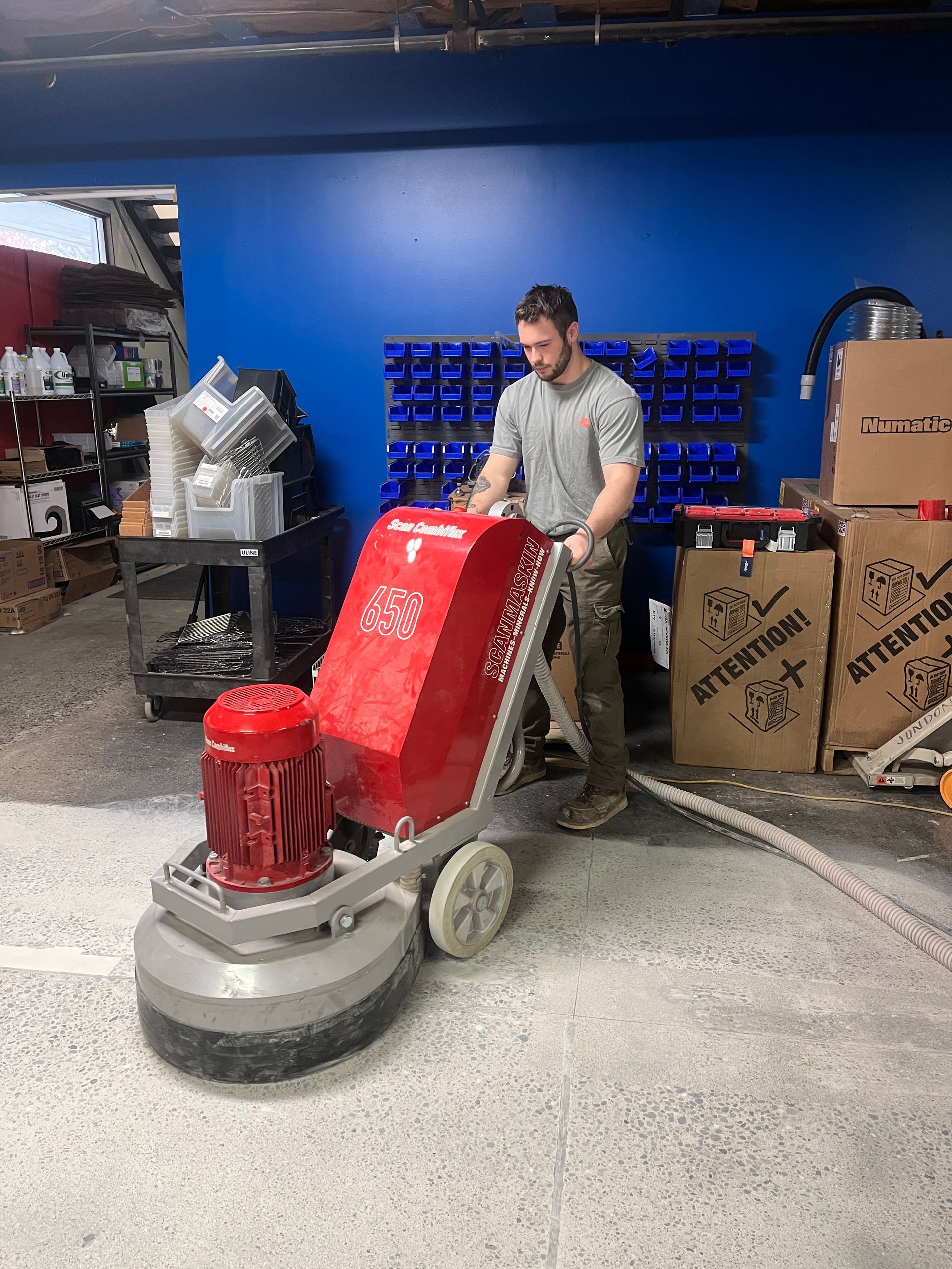 A man operates a large red floor grinder on a speckled concrete surface. He's in a warehouse with blue walls and shelves.