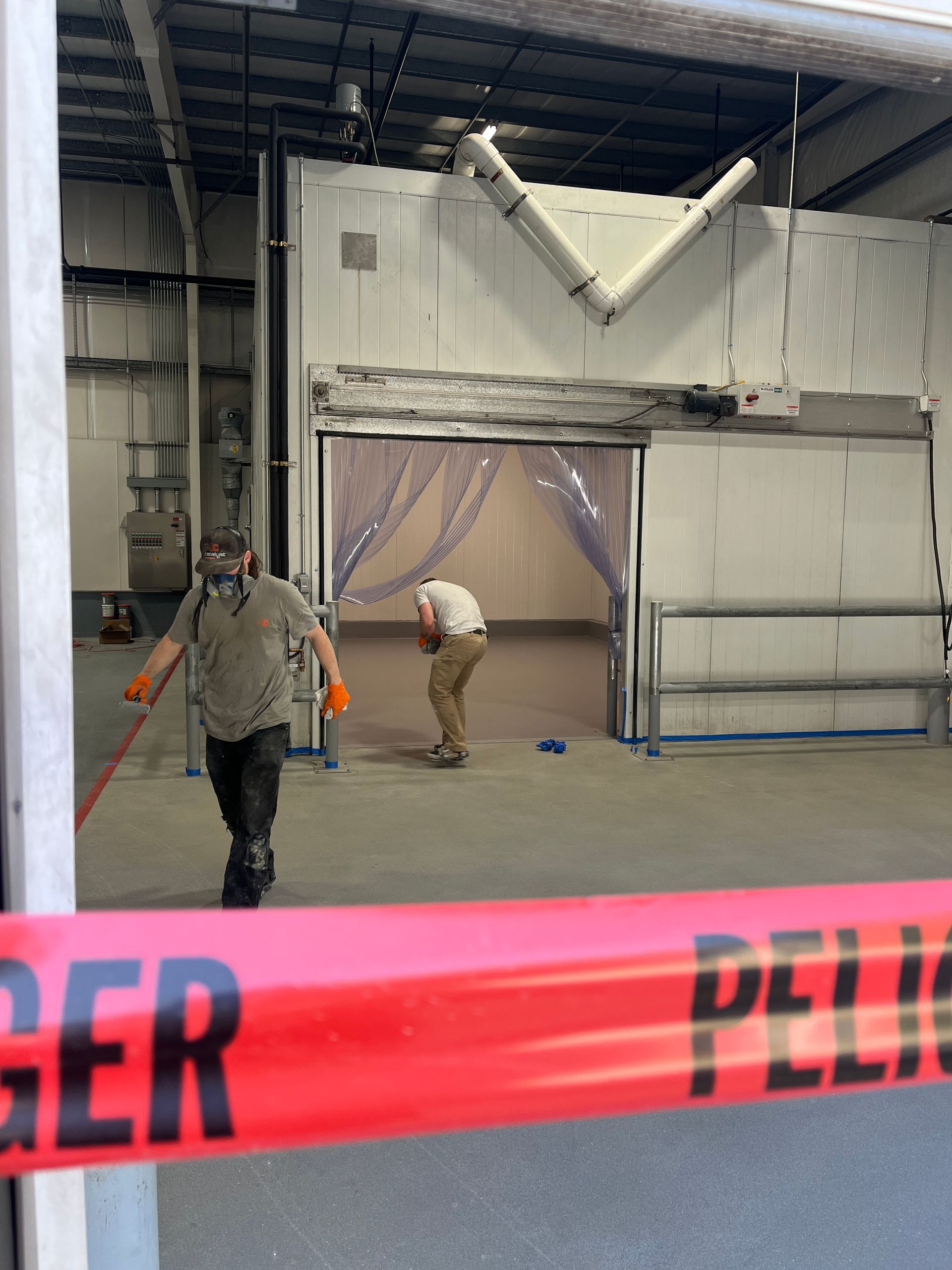 Red caution tape across foreground. Two men, one wearing a respirator, in an industrial building, possibly inspecting equipment.