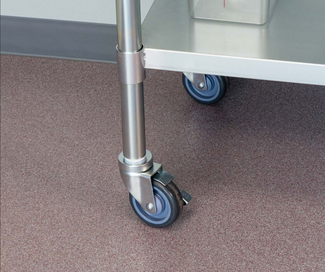 Close-up of a stainless steel cart with blue wheels on a brown speckled floor. The wheels have a locking mechanism.