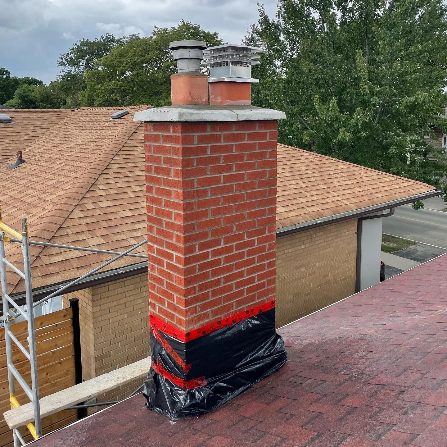 Brick chimney on a red roof, wrapped in black plastic, with metal chimney caps.