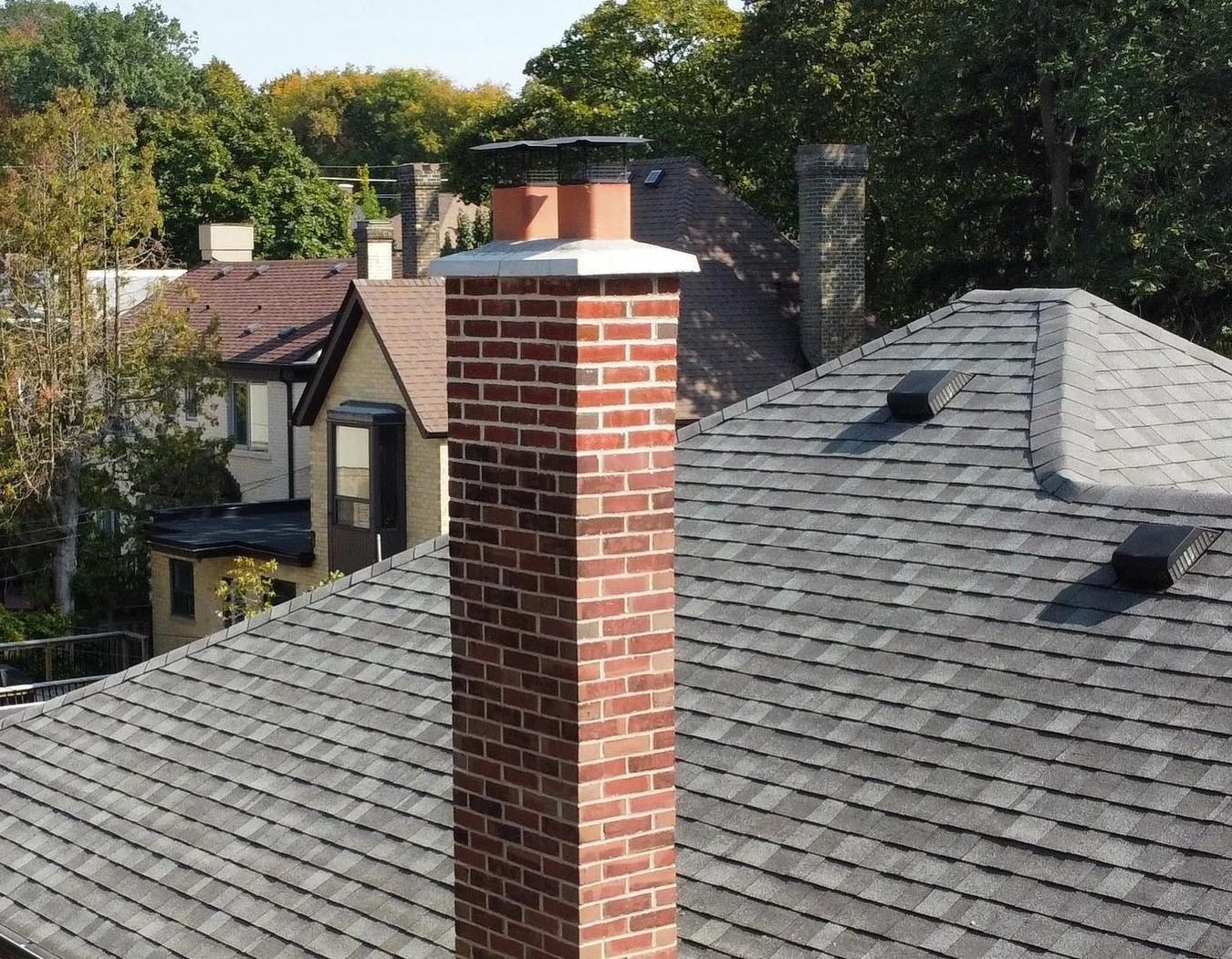 Brick chimney on a gray shingle roof, other houses and trees in background on a sunny day.