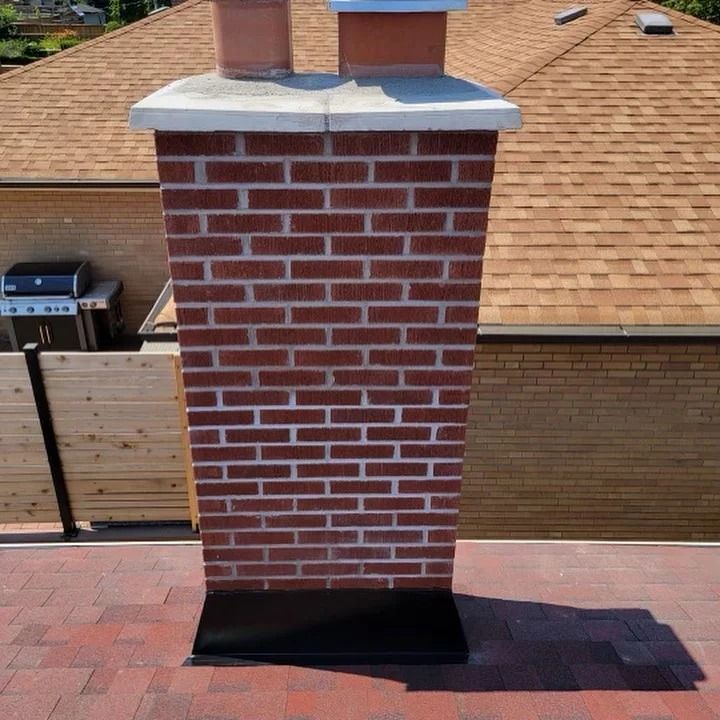 Brick chimney on a brown shingled roof with a black metal base and two flue caps.