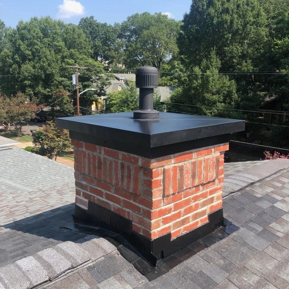 Brick chimney with black cap and vent on a rooftop, surrounded by trees.