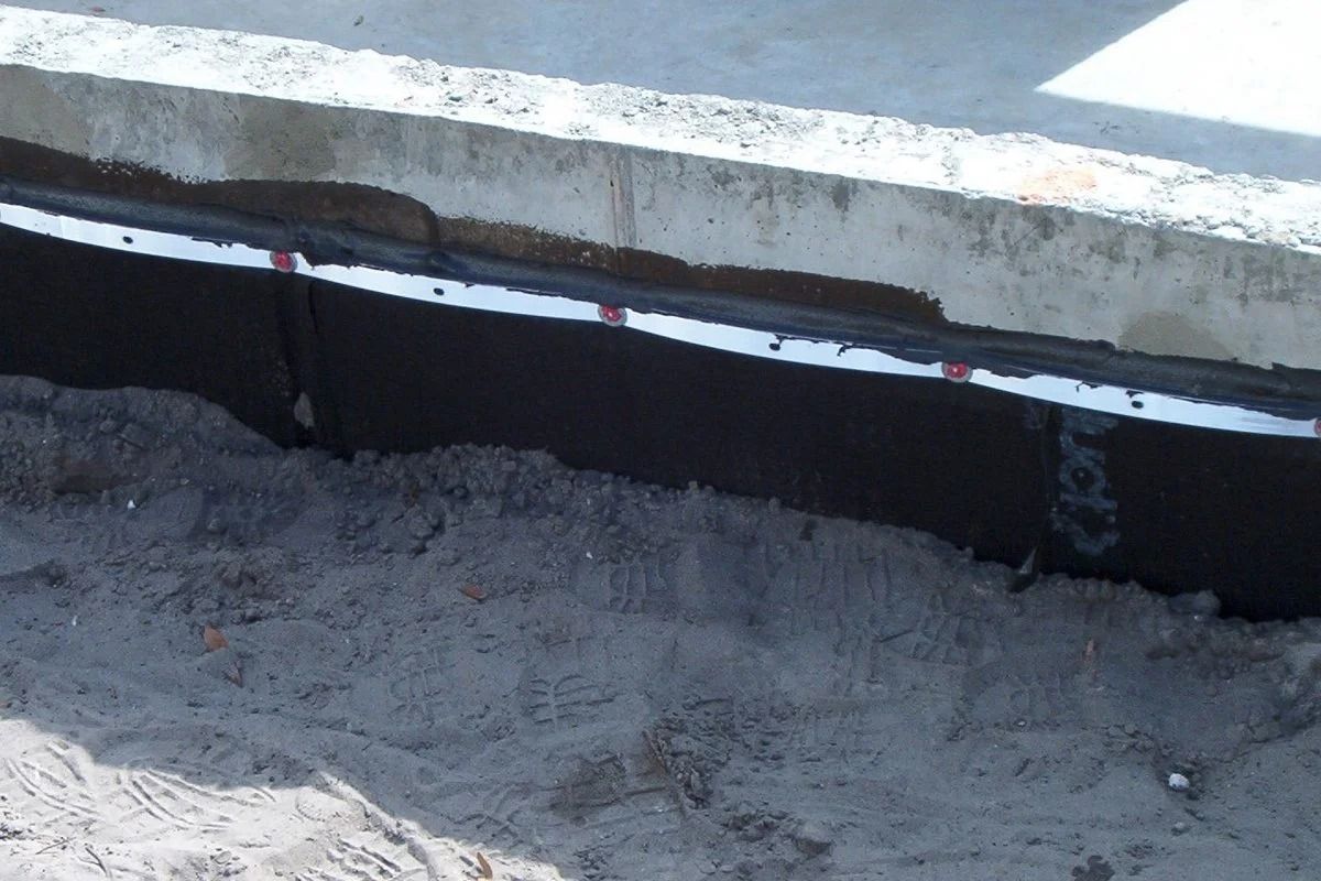 Black waterproofing layer along concrete foundation with white pipe.