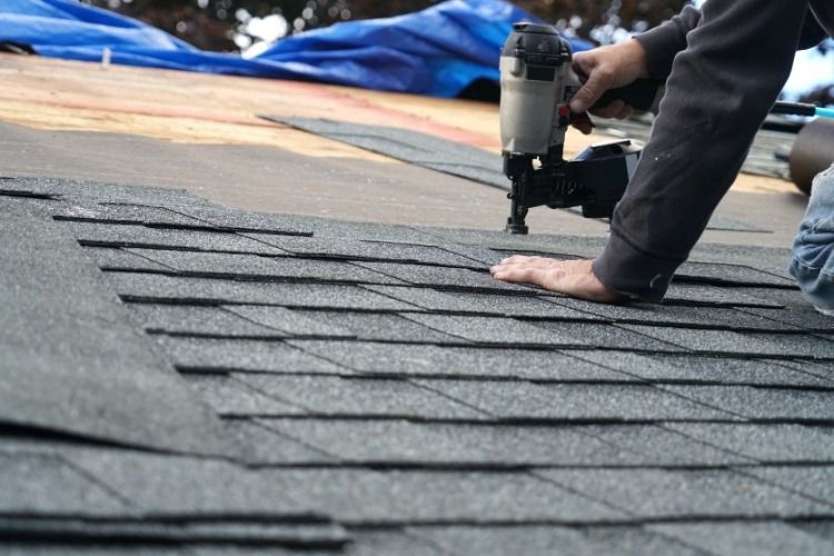 Person using a nail gun to install gray shingles on a roof.