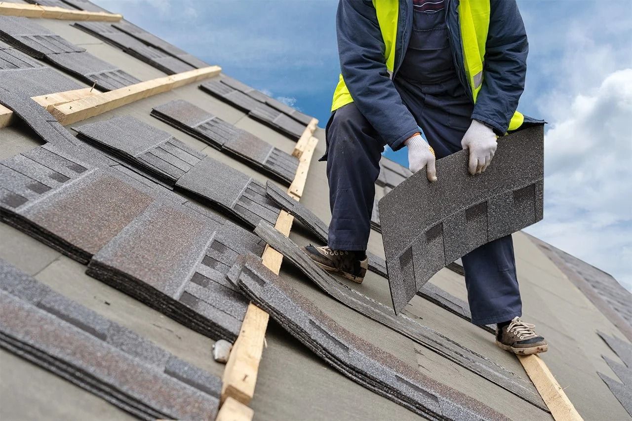 Roofer installing asphalt shingle on roof, wearing a yellow safety vest and gloves.