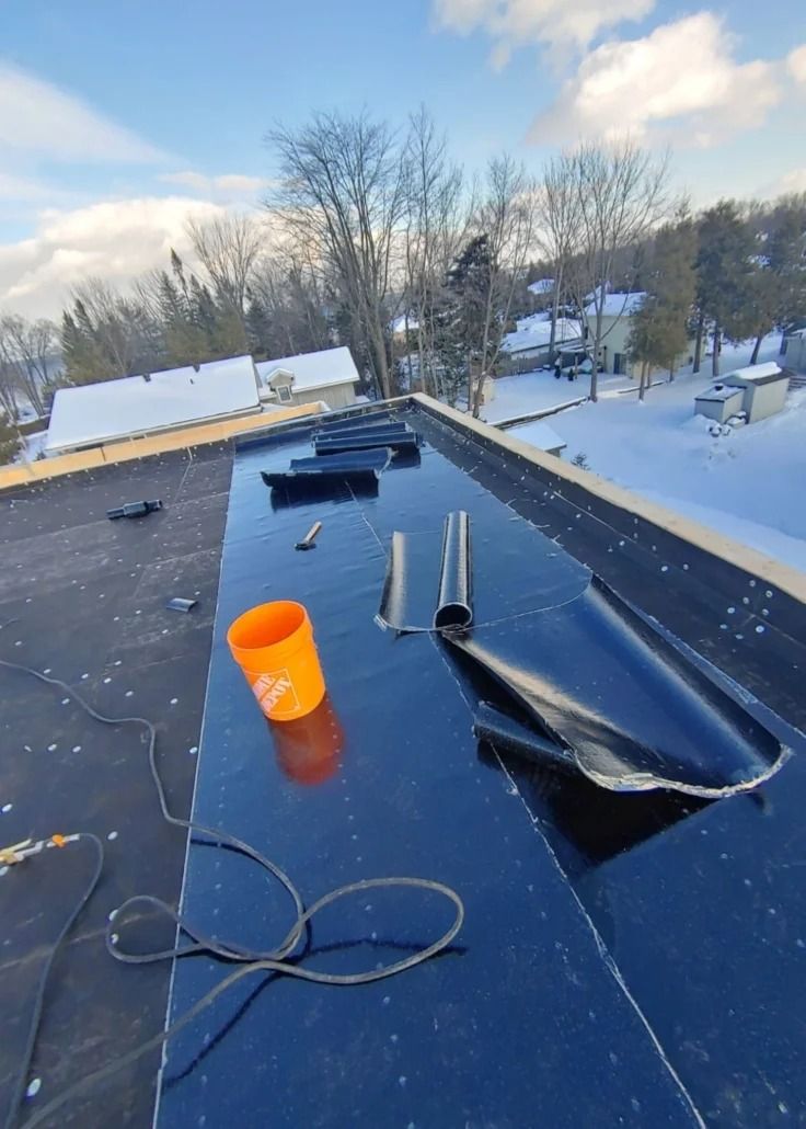 Black roofing material with tools and an orange cup on a roof covered in snow, in a winter setting.