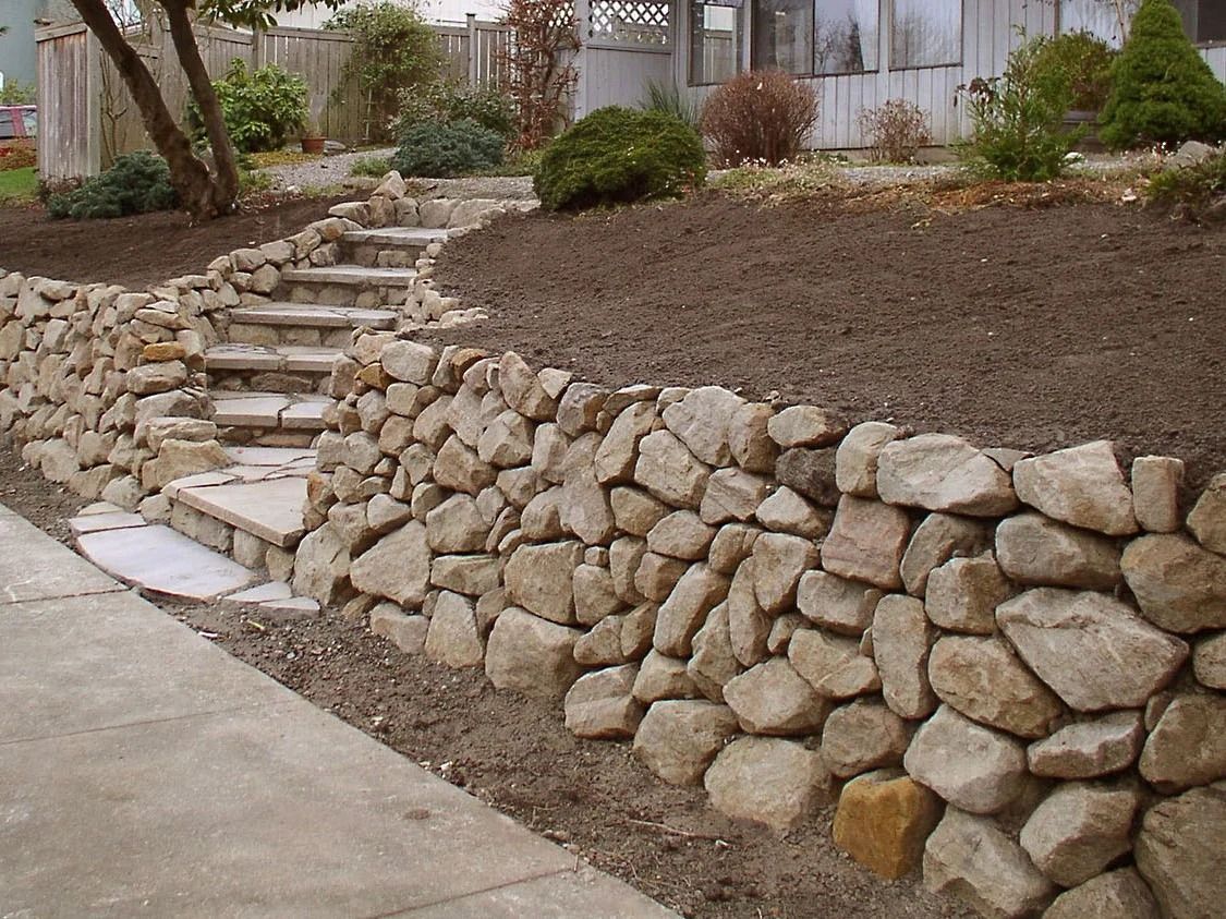 Stone retaining wall with steps leading up to a landscaped area with plants and trees.