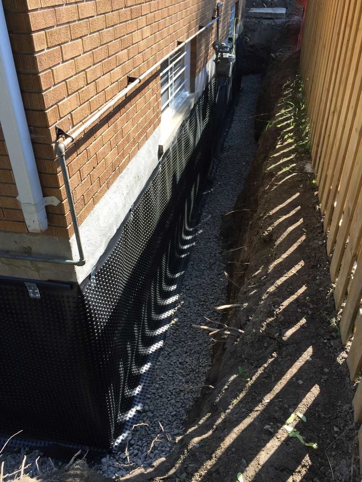 Foundation repair: Trench next to a brick house with black drainage membrane and gravel.