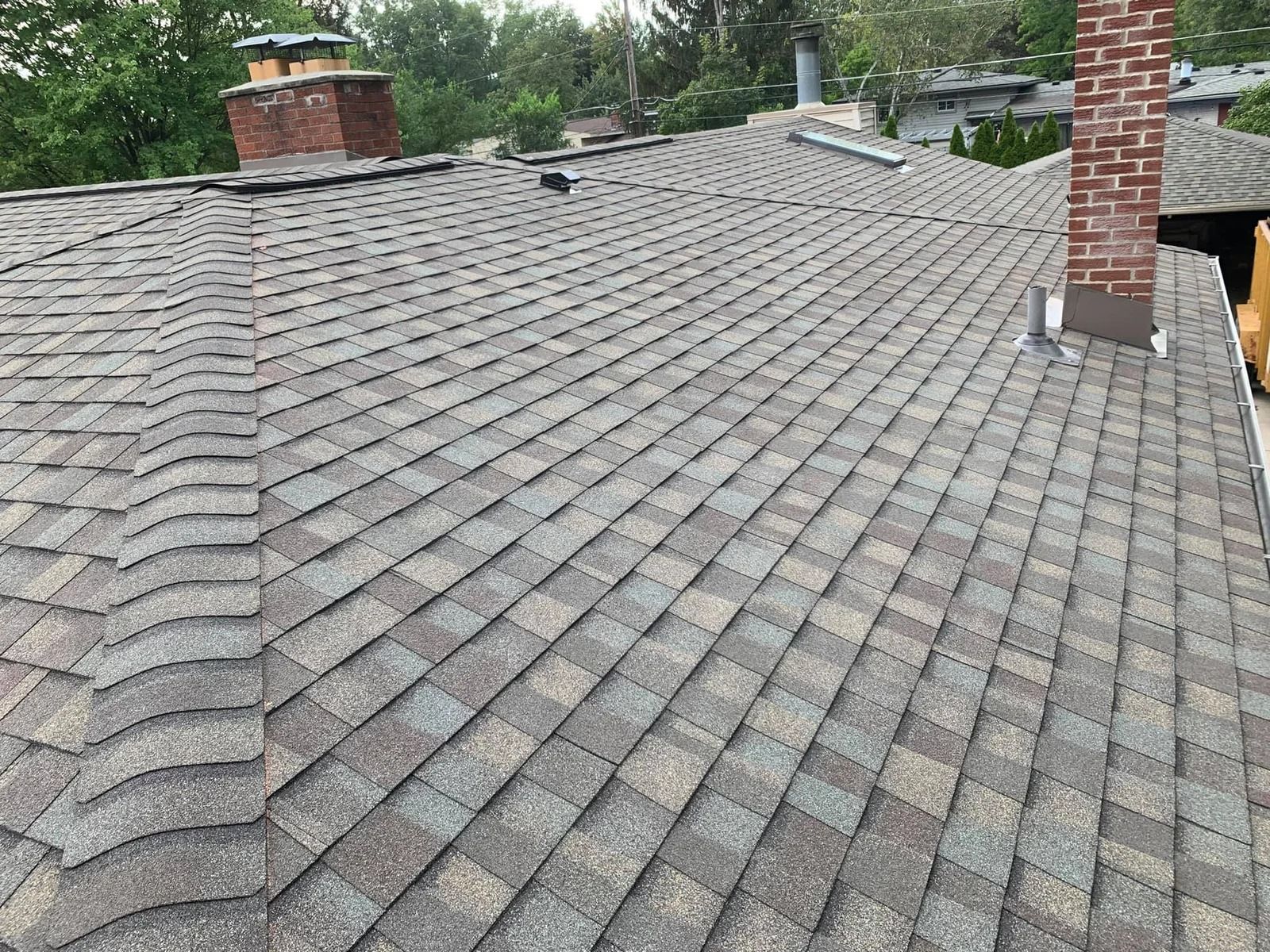 A multi-toned asphalt shingle roof with two brick chimneys against a tree-lined background.