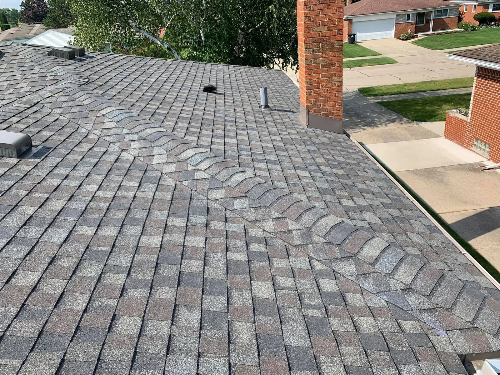 View of a roof with damaged shingles near a brick chimney and residential setting.