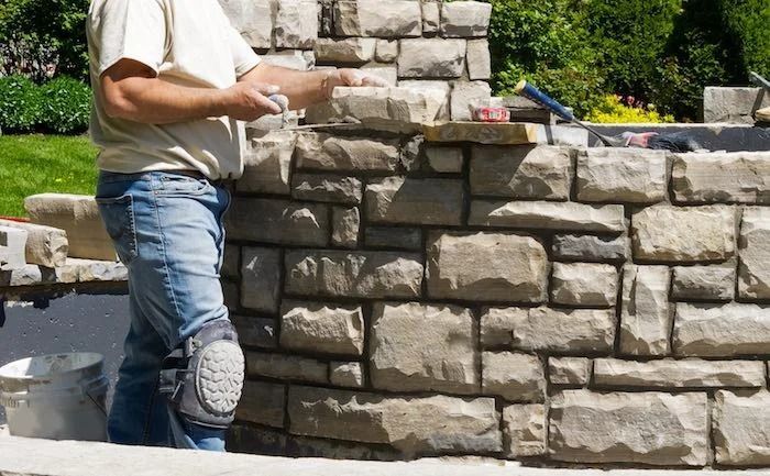 Man laying stone bricks, wearing knee pads, building a stone wall outdoors.
