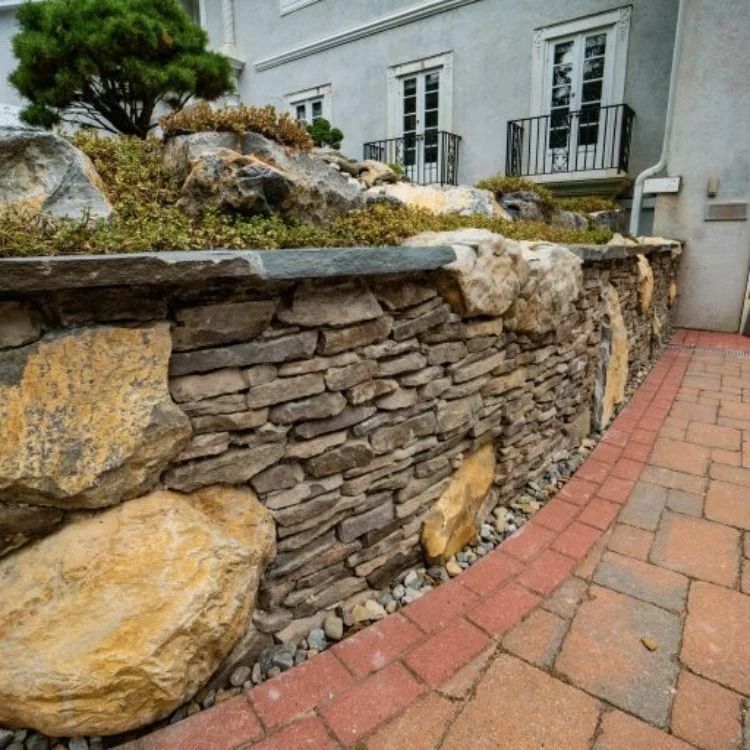 Stone retaining wall with large boulders, topped by a bed of small plants and rocks, beside a brick pathway.