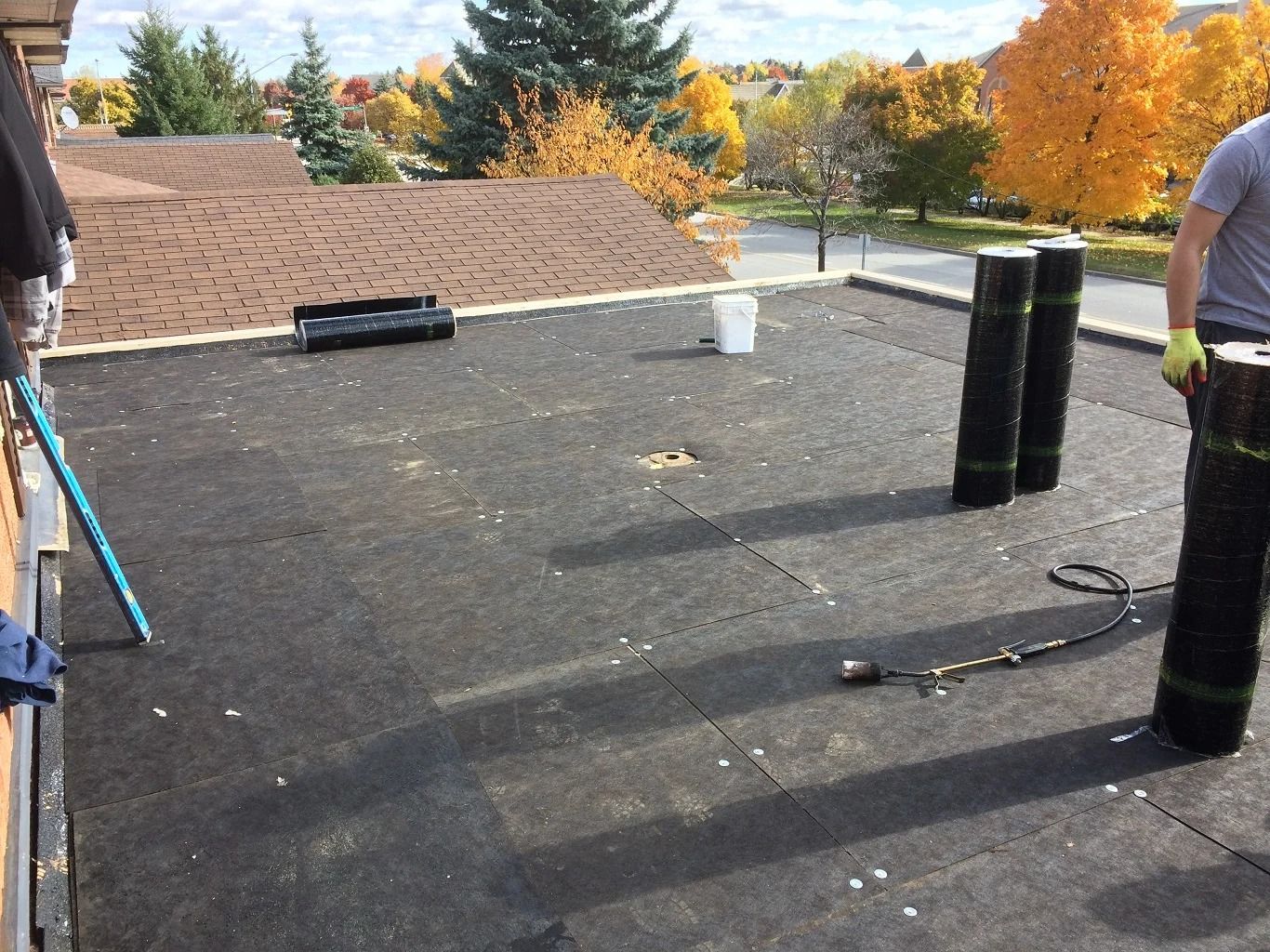 Flat roof with rolled roofing material and worker holding a roll. Autumn trees in background.