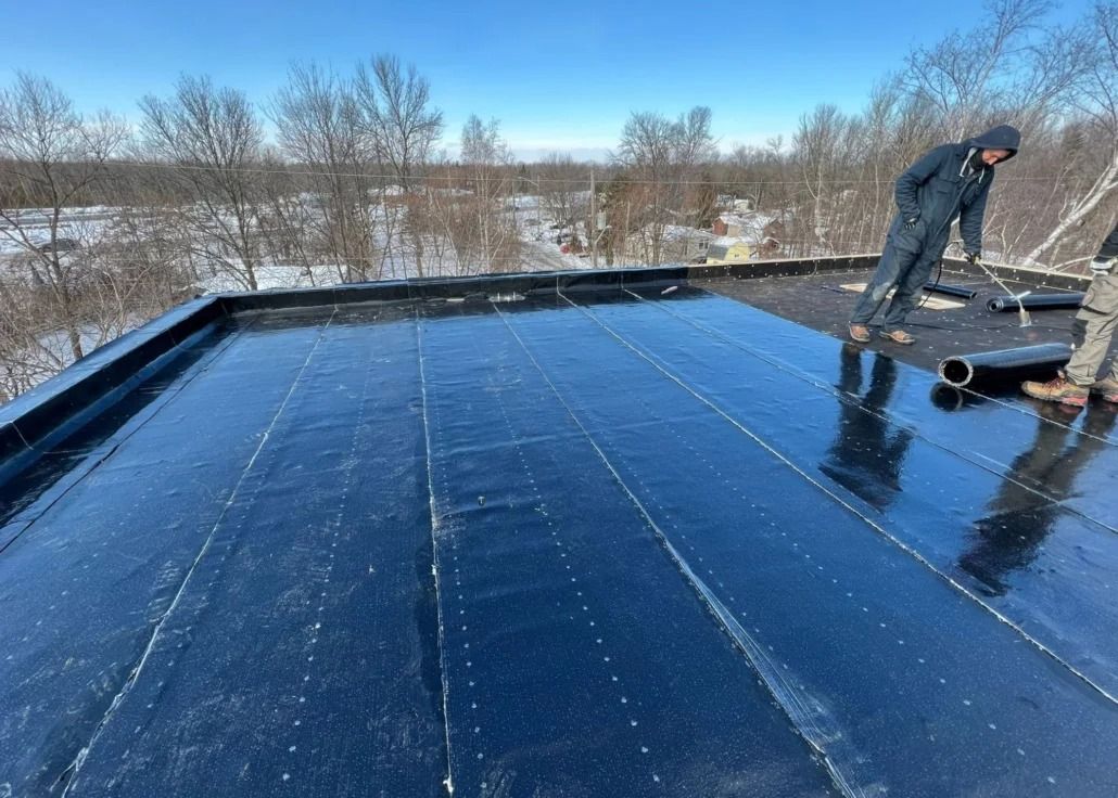 Workers installing black roofing material on a flat roof, snowy background.