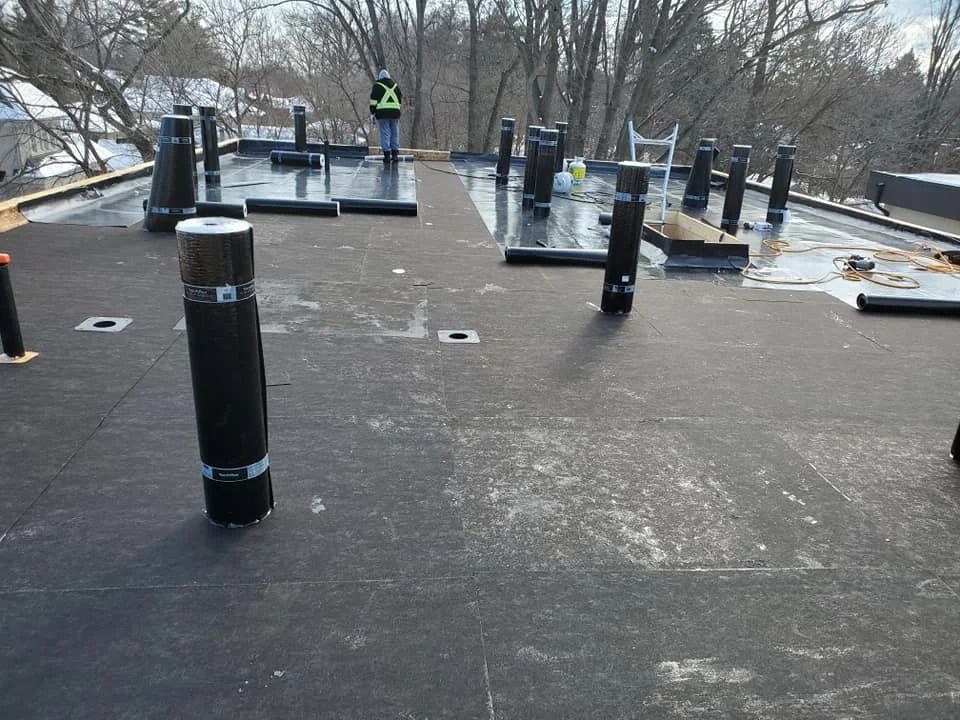 Black roofing material being installed on a roof with vertical pipes, two workers in the background.