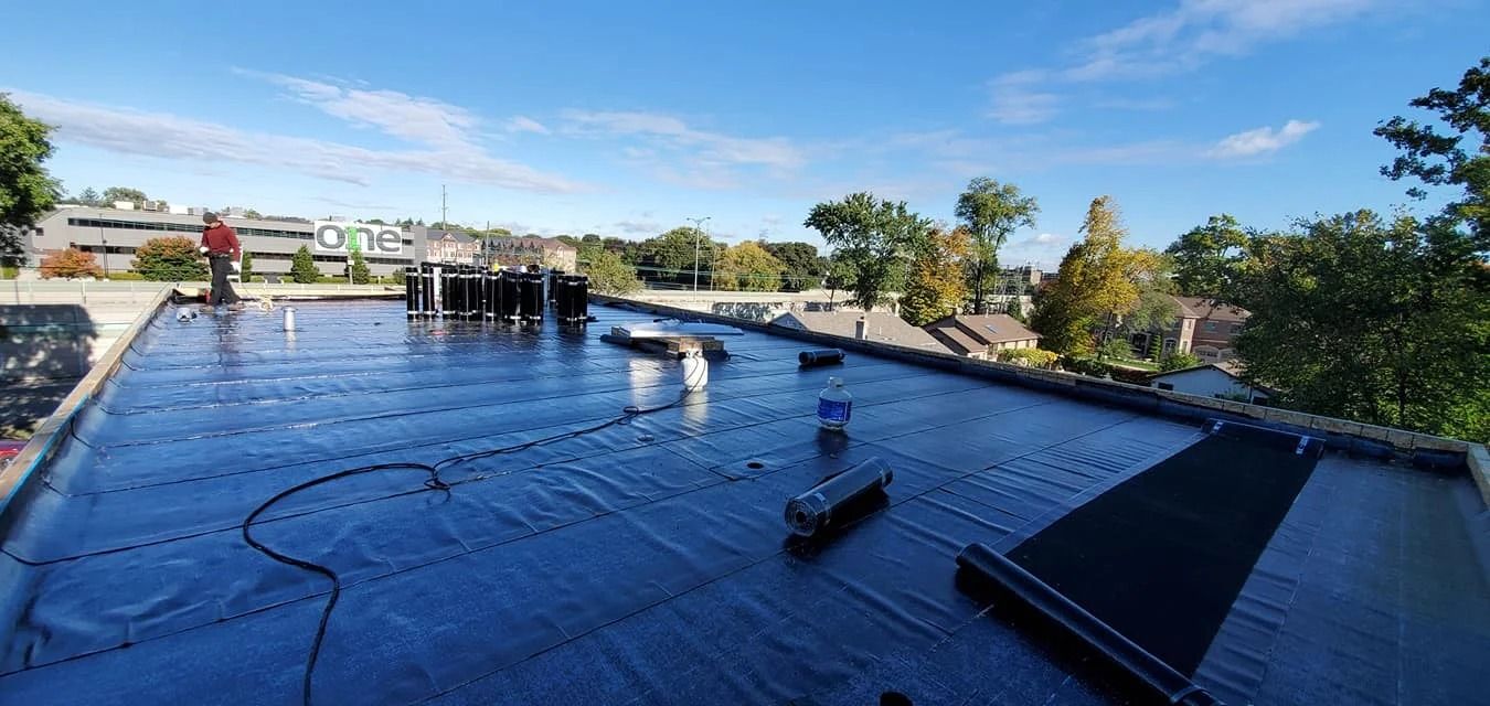 A flat roof with a new black membrane being installed, with buildings and trees in the background under a blue sky.