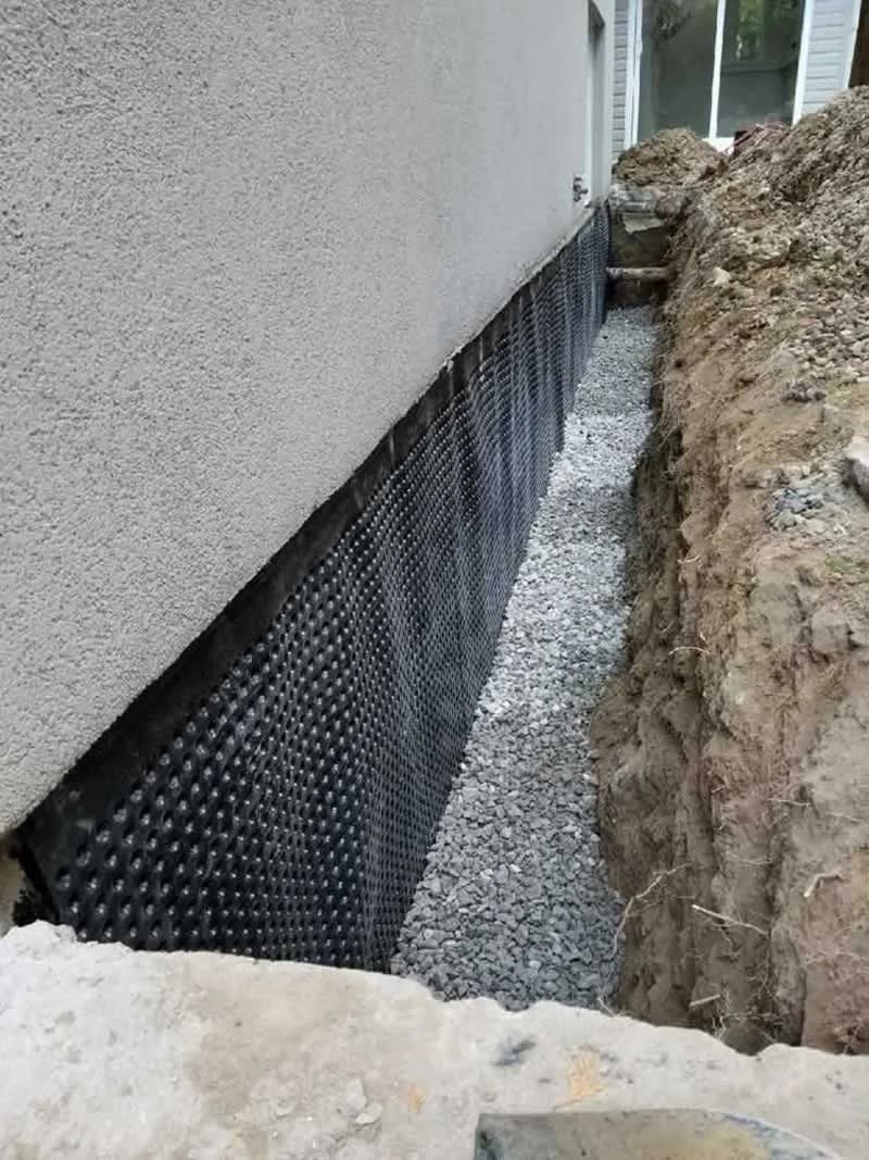 Foundation waterproofing; black dimpled membrane against a building wall, gravel in a trench.