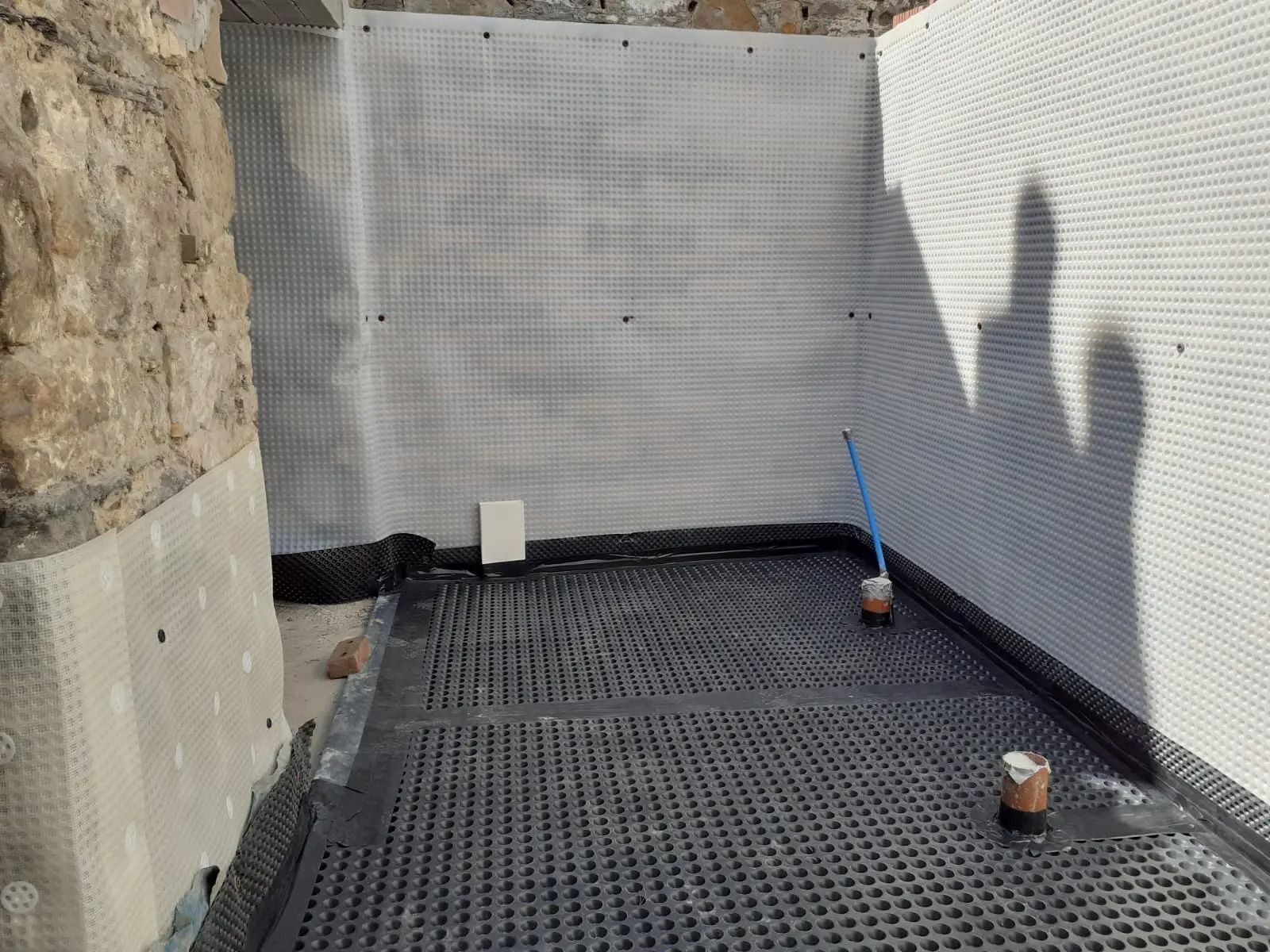 Basement waterproofing: perforated black membrane on floor, white mesh on walls, against a stone wall.