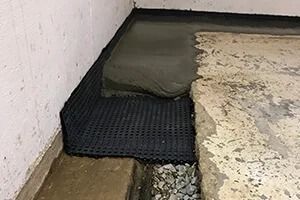 Black drainage mat and wet cement against a concrete wall and floor, part of a foundation repair.