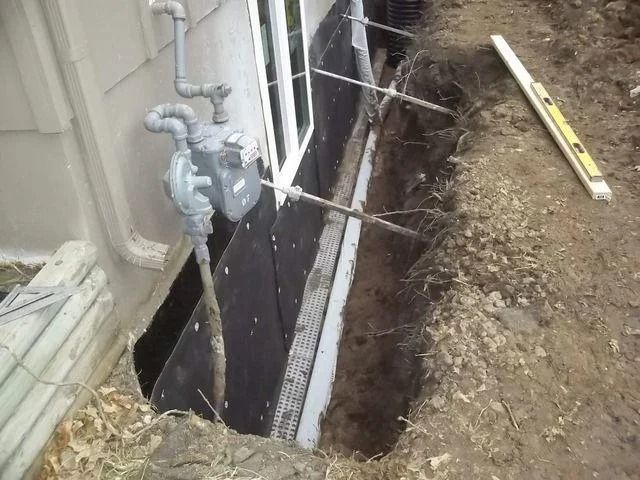 Construction site near a house's foundation. A trench reveals black waterproofing, a window, and gas meter. 