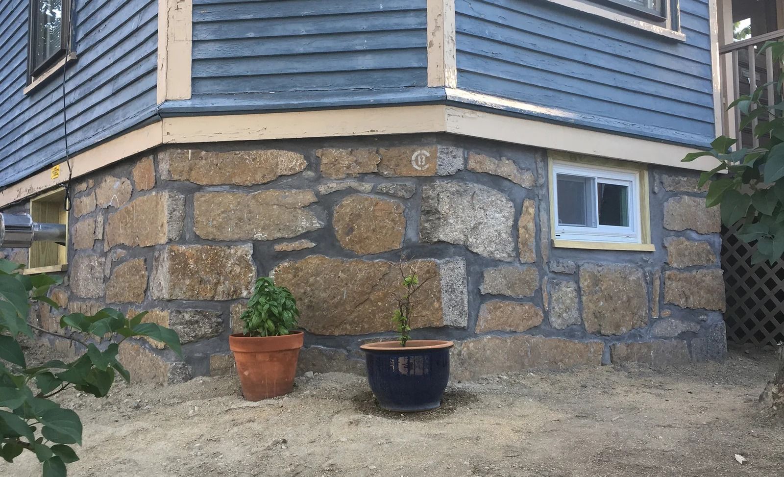 Corner of a house with blue siding, a stone foundation, and potted plants.