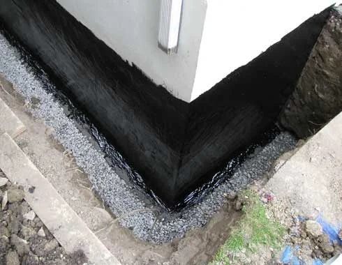 Black waterproof membrane applied to foundation corner, surrounded by gravel and earth.