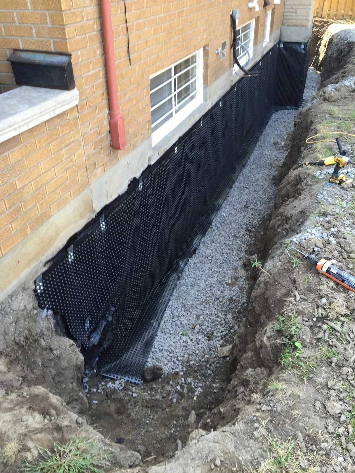 Foundation waterproofing with black drainage membrane and gravel in a trench next to a brick building.