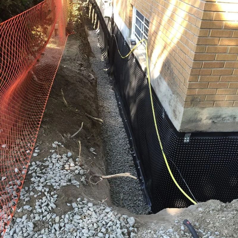 Excavation along a brick building, revealing foundation drainage and gravel, with orange safety fencing.