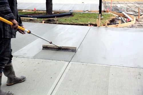 Person using a concrete brush on a sidewalk, smoothing wet cement.