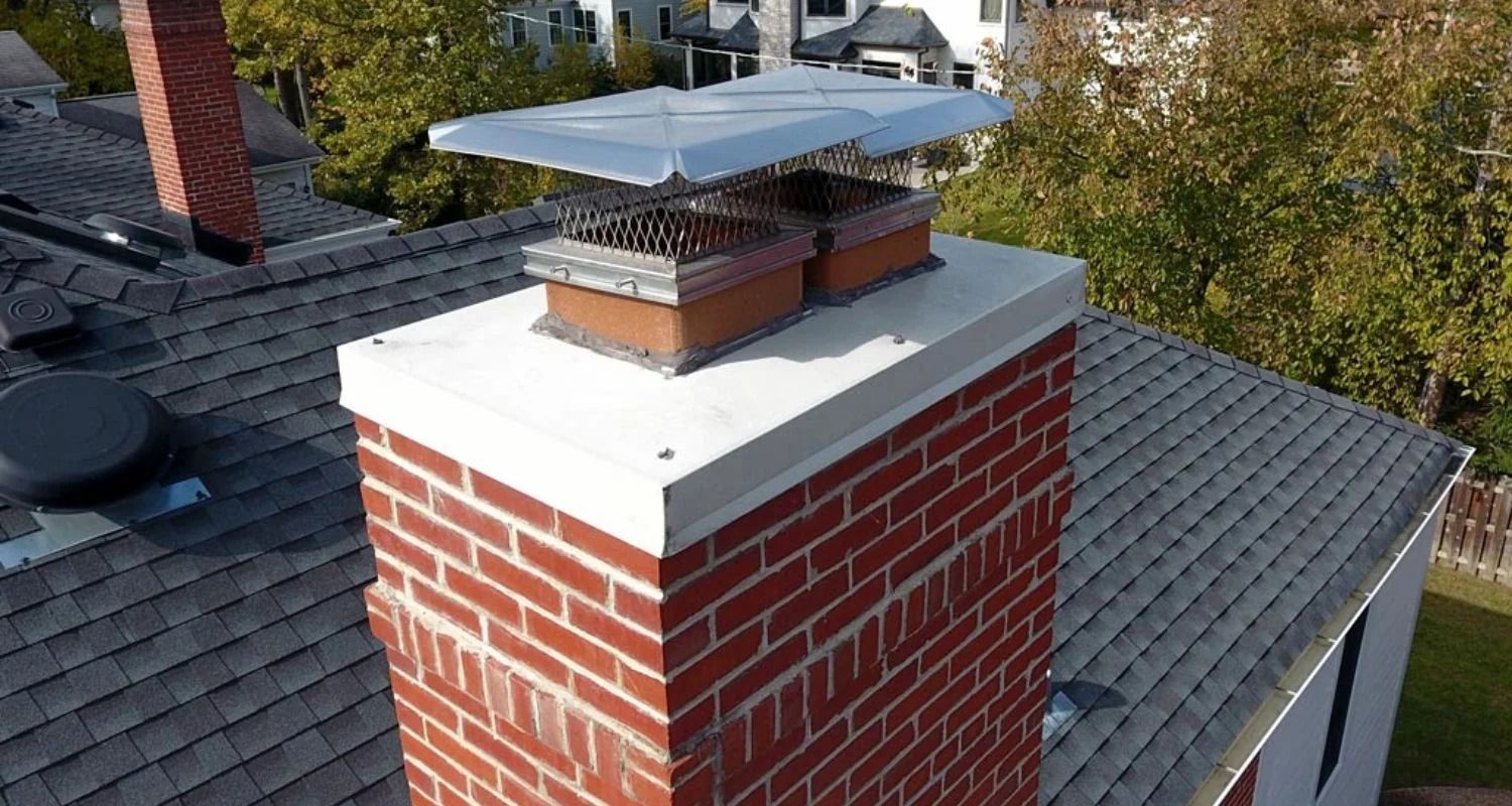 Brick chimney on a roof with a metal cap and mesh, next to trees and houses.