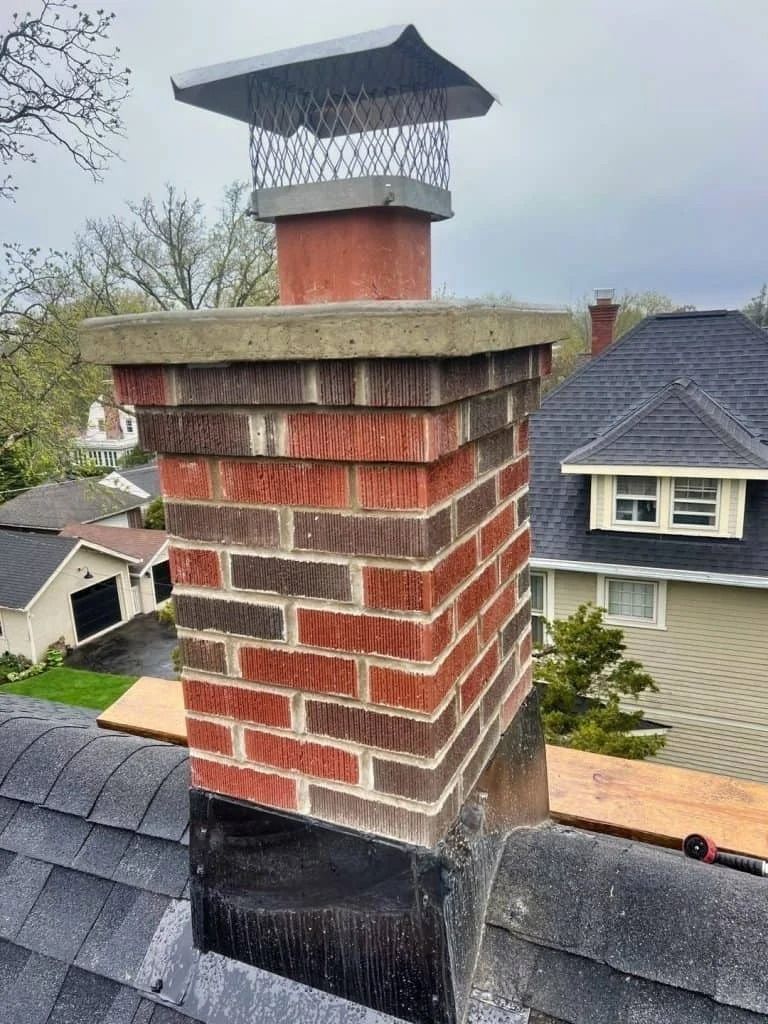 Brick chimney on a roof with a metal cap, red and brown bricks, gray concrete cap, and a cloudy sky.