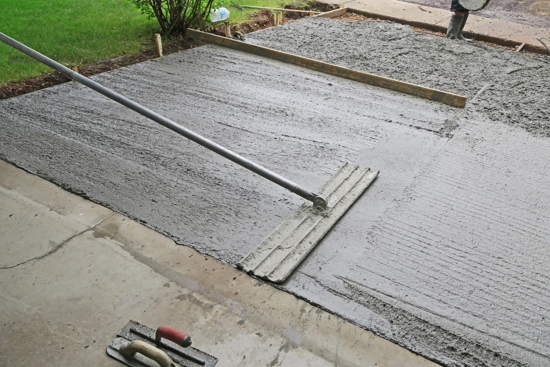 Person using a concrete rake to level wet concrete in a driveway, surrounded by borders and tools.