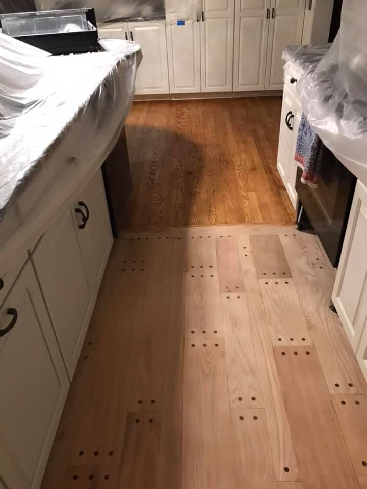 Kitchen with light wood floor being installed, contrasting with existing dark wood. Cabinets covered in plastic.