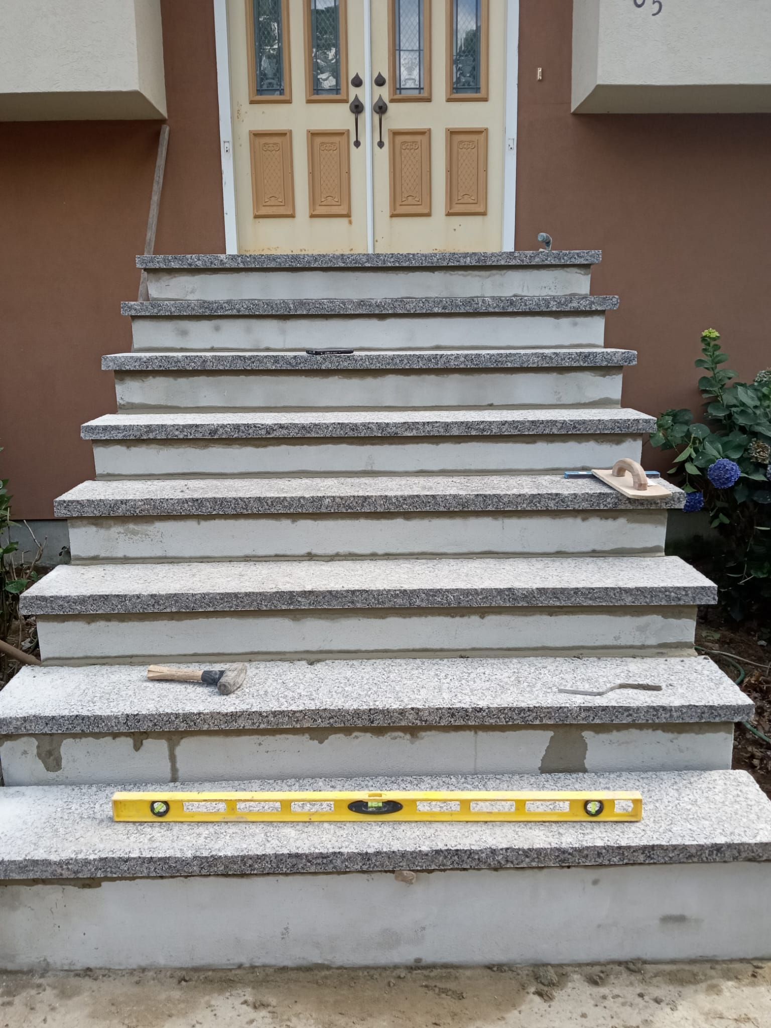 Concrete front steps of a house, a level rests on the bottom step. Doors are in the background.