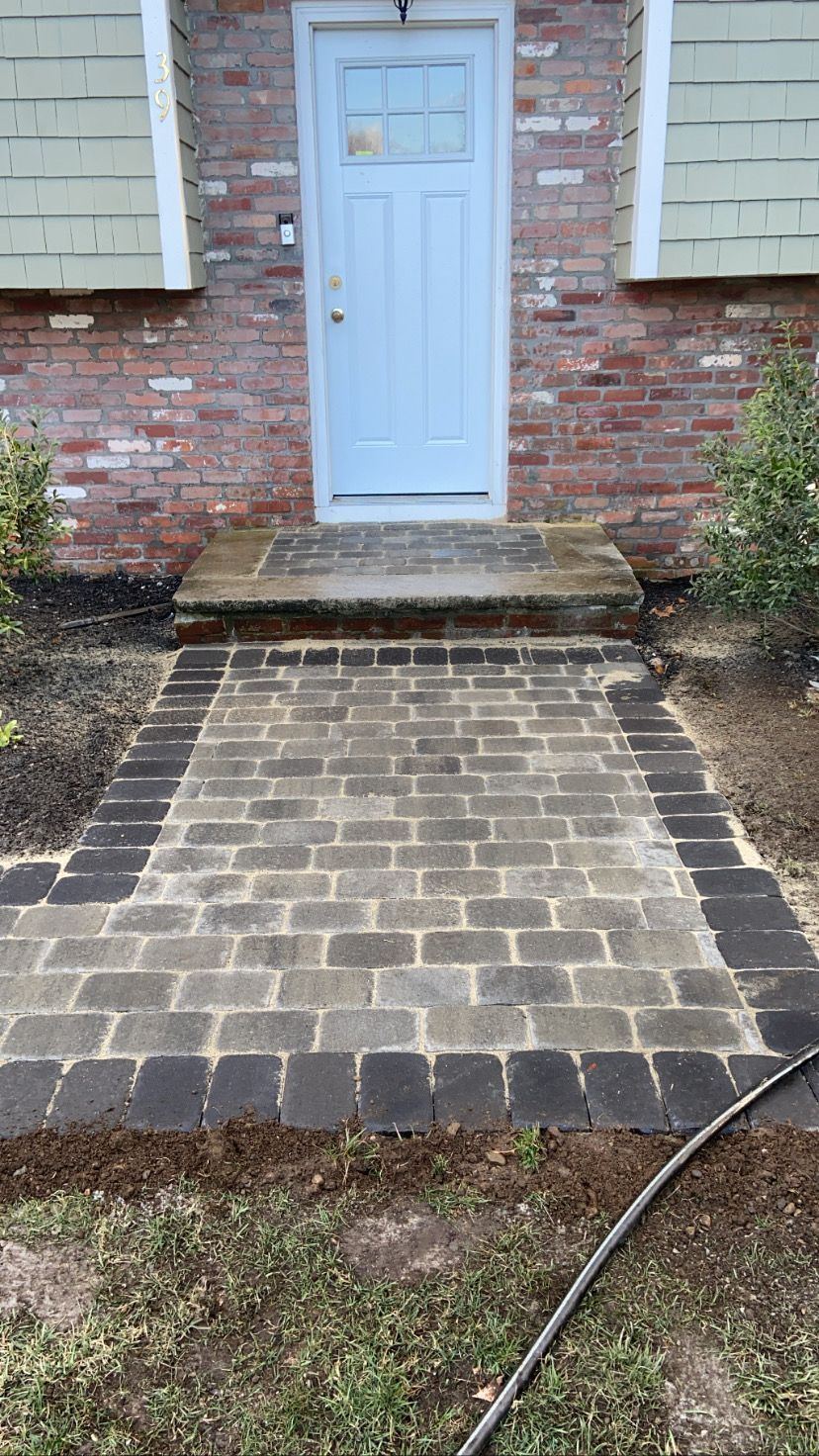 Brick walkway leading to a light blue door, bordered by darker bricks and flanked by brick and shrubbery.