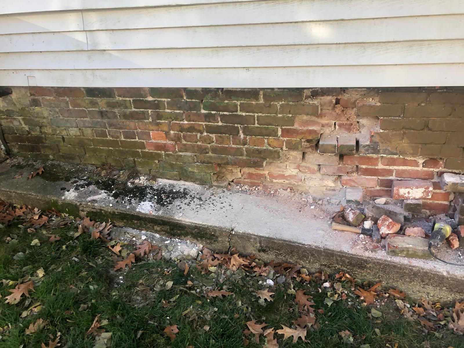 Damaged brick foundation of a house, with crumbled bricks, white siding, and green grass.