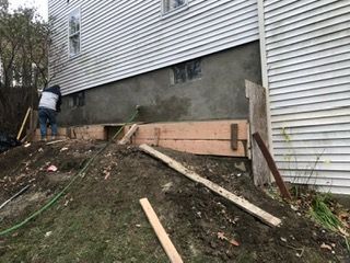 Construction worker forming a concrete foundation against a house. Earth mound in front, wood forms in place.